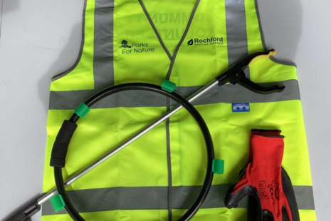 Photo shows a high vis jacket in yellow, laying on top of that is some reg protection gloves, a hoop that holds the bag for rubbish and a litter grabber.